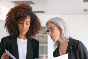 Content young manager showing document to adult colleague. Two pretty content female colleagues holding papers and standing in office room. Teamwork, business and management concept